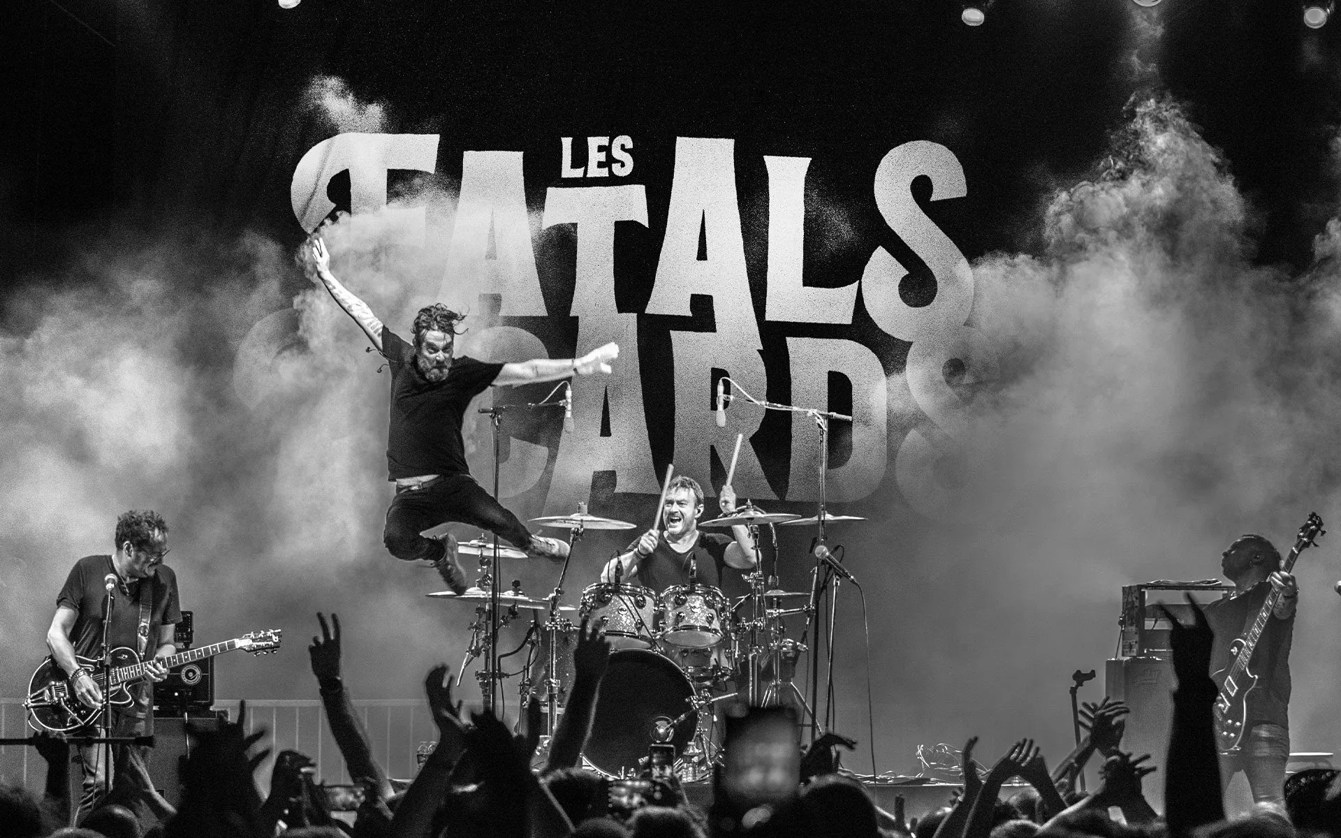 Les Fatals Picards en plein concert : saut du chanteur devant la batterie de Jean-Marc, logo du groupe dans la fumée